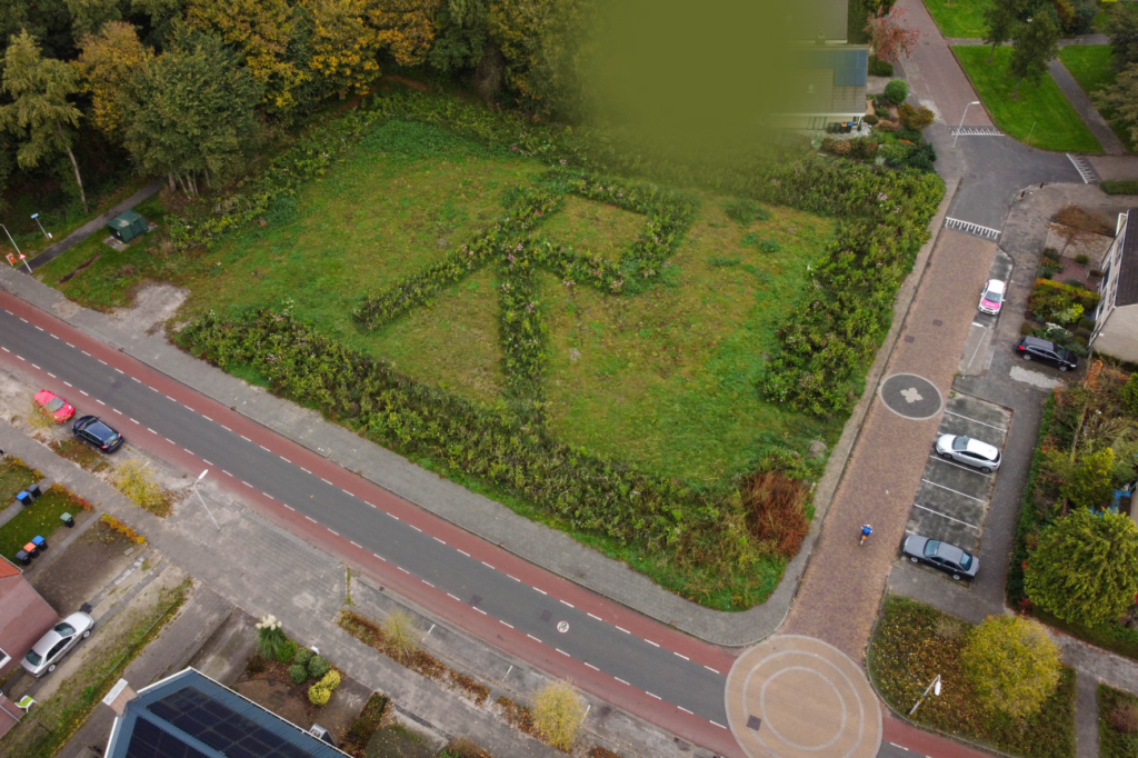 Drone foto van Buitenom 1 in Rutten. Een grasveld omgeven door bos een straten. Het grasveld is omzoomt met bossage en met planten is een grote letter R gemarkeerd in het grasveld.